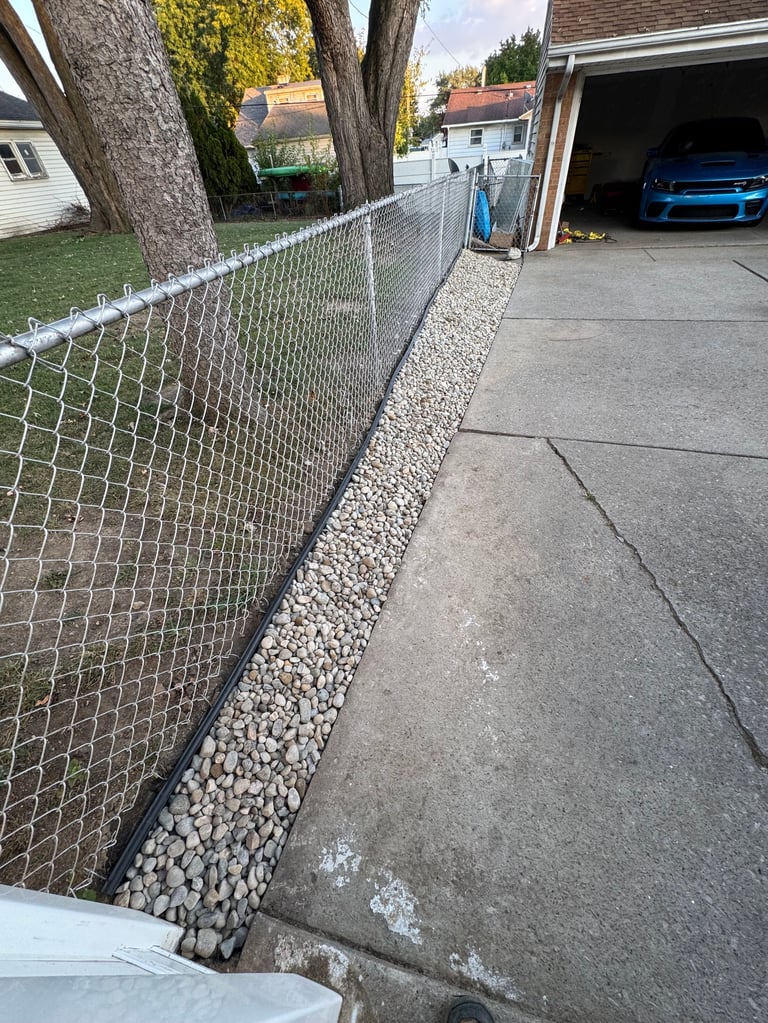 Chain-link fence along driveway with gravel base, residential garage with blue car visible, tree trunk and green lawn