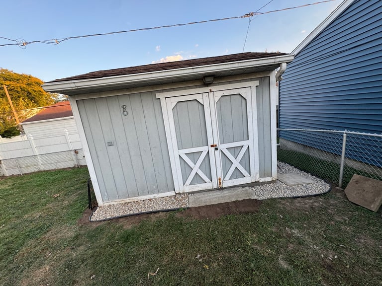 White wooden shed with double doors and X-pattern design on a residential property with grass and fencing